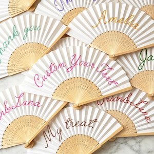May include: A collection of white folding hand fans with light wood frames. Each fan features custom text in various colors and cursive fonts, including phrases like Thank you, Amelia, Jasper, Custom Your Text, Orion & Luna, and My treat.