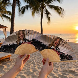 May include: Two open hand fans with a photo of a person and the text "Caitlin's 18th Birthday". The fans have a natural wood handle and a printed image. The background shows a beach scene with palm trees and a sunset.