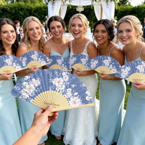 May include: A handheld folding fan featuring a dusty blue fabric canopy printed with a white and pink floral pattern. The wooden ribs are light-colored, and the text "Dean & Laurel" is printed in a script font on the lower portion of the fan.