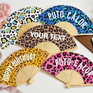 Op de afbeelding: Een verzameling handwaaiers met luipaardprint in verschillende kleuren, waaronder blauw, bruin, geel en roze. Sommige waaiers hebben tekst zoals "HAPPY BIRTHDAY", "YOUR TEXT" en "PUTO CALOR". De waaiers zijn gemaakt van hout en stof.