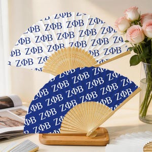 May include: Two folding fans with wooden handles. One fan is navy blue with white text, and the other is white with navy blue text. The fans are displayed on a wooden tray with a vase of pink roses in the background.