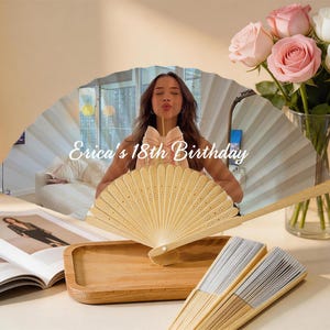 May include: A decorative hand fan with a light wood handle and a cream-colored fan. The fan is displayed with a wooden tray, open magazines, and a bouquet of pink and white roses. The text "Erica's 18th Birthday" is visible.