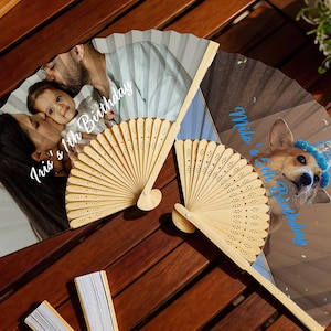 May include: Two personalized hand fans with wooden handles. One fan features a family photo with the text "Iris's 1th Birthday". The other fan displays a dog wearing a party hat with the text "Milo's 1th Birthday".