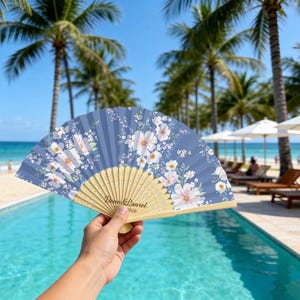 May include: A folding hand fan with a dusty blue fabric canopy featuring a white and light pink floral pattern. The fan has light-colored wooden ribs and the text Dean & Laurel 2026 printed in black on the lower portion of the wooden frame.