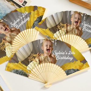 May include: Hand fans with a photo of a baby girl, the text "Sylvie's 2th Birthday", and "Happy Birthday" on a gold cake topper. The fans have a light wood handle and a gray, yellow, and white design.