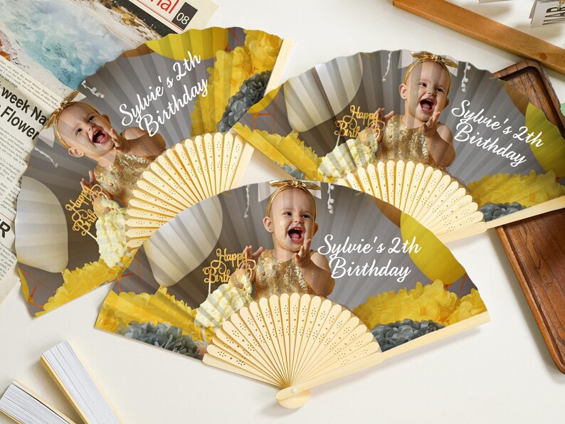May include: Hand fans with a photo of a baby girl, the text "Sylvie's 2th Birthday", and "Happy Birthday" on a gold cake topper. The fans have a light wood handle and a gray, yellow, and white design.