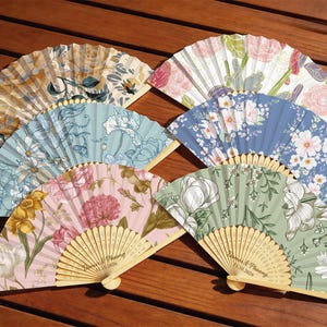 May include: Six folding hand fans with wooden ribs and fabric leaves featuring various floral patterns in shades of blue, pink, green, and yellow. Two fans display the text "Dames & Phuong 13.10.2026" engraved on the wooden handle section.