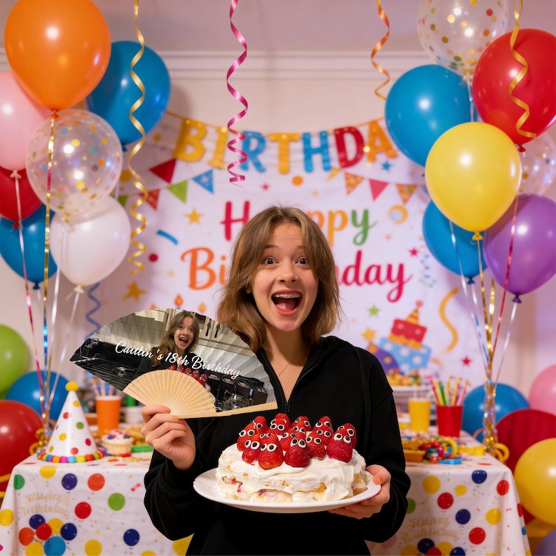 May include: A young person smiles, holding a decorated cake and a fan with a photo and text. The cake is topped with strawberries. Colorful balloons and a birthday banner are in the background. The fan reads "Caitlin's 18th Birthday."