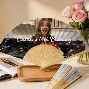 May include: A decorative fan with the text "Caitlin's 18th Birthday" is displayed. The fan is made of light-colored wood and is partially open. A bouquet of pink and white roses sits in a glass vase. A wooden tray and open magazine are also present.