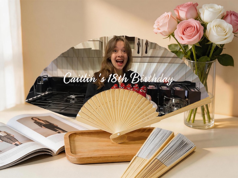 May include: A decorative fan with the text "Caitlin's 18th Birthday" is displayed. The fan is made of light-colored wood and is partially open. A bouquet of pink and white roses sits in a glass vase. A wooden tray and open magazine are also present.