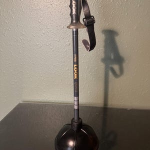 Puede incluir: Un bastón de esquí negro y gris con un mango y una correa de muñeca negros. El bastón tiene el texto "LOOK 9000" impreso. El bastón está unido a un desatascador de goma negro.