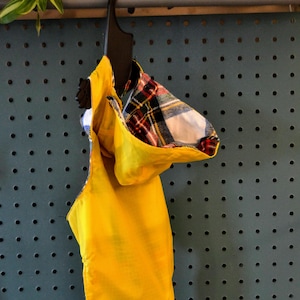 Op de afbeelding: Een gele regenjas met capuchon en een geruite voering. De regenjas hangt aan een hanger tegen een blauwe prikbord.