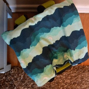 May include: A blue and green patterned fabric with a white background. The fabric has a wavy design that resembles clouds. The fabric is folded and draped over a chair.
