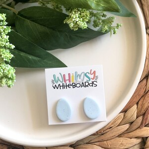 May include: A pair of light blue oval-shaped earrings with a speckled texture. The earrings are on a white card with the text "Whimsy Whiteboards" printed on it.