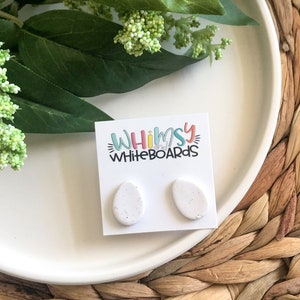 May include: A pair of white oval-shaped earrings with a speckled texture. The earrings are displayed on a white card with the text "Whimsy Whiteboards".