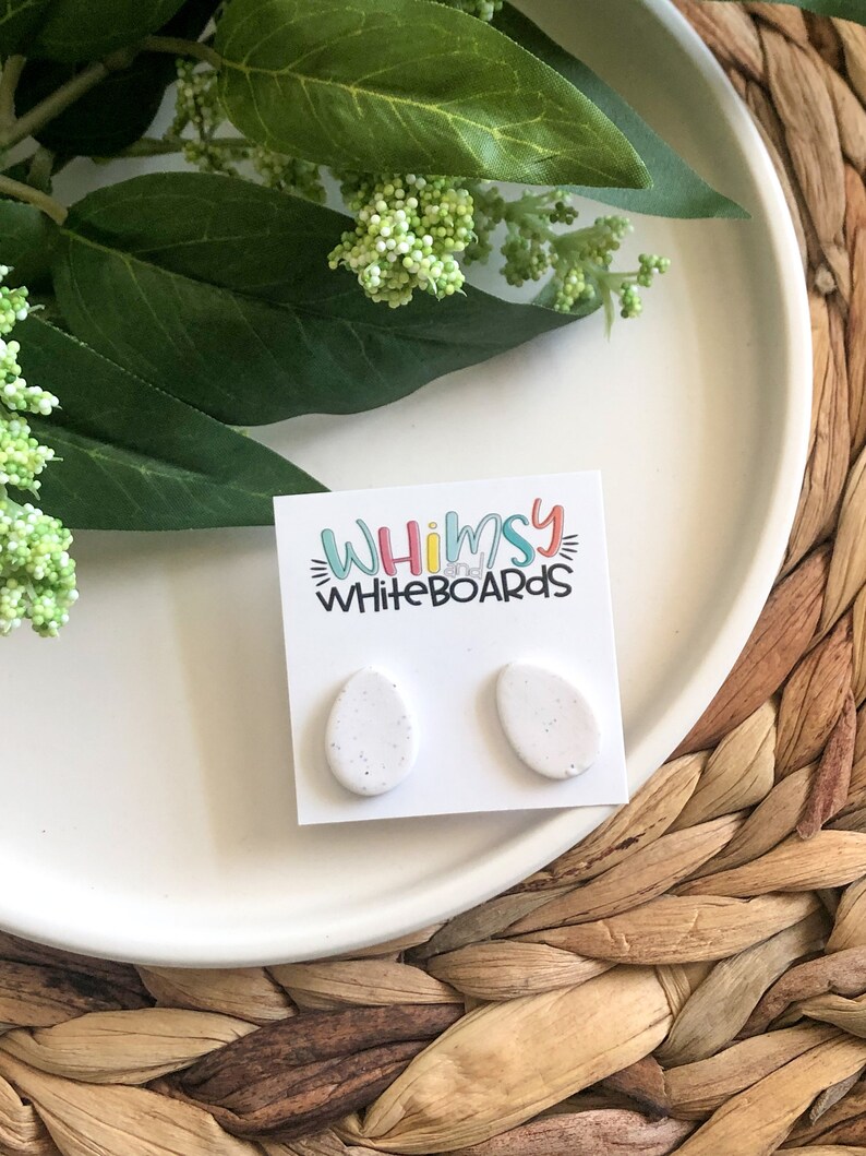 May include: A pair of white oval-shaped earrings with a speckled texture. The earrings are displayed on a white card with the text "Whimsy Whiteboards".