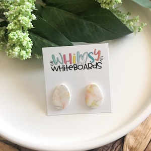 May include: A pair of white oval-shaped earrings with a marbled design in shades of pink, yellow, and green. The earrings are displayed on a white card with the text "Whimsy Whiteboards".