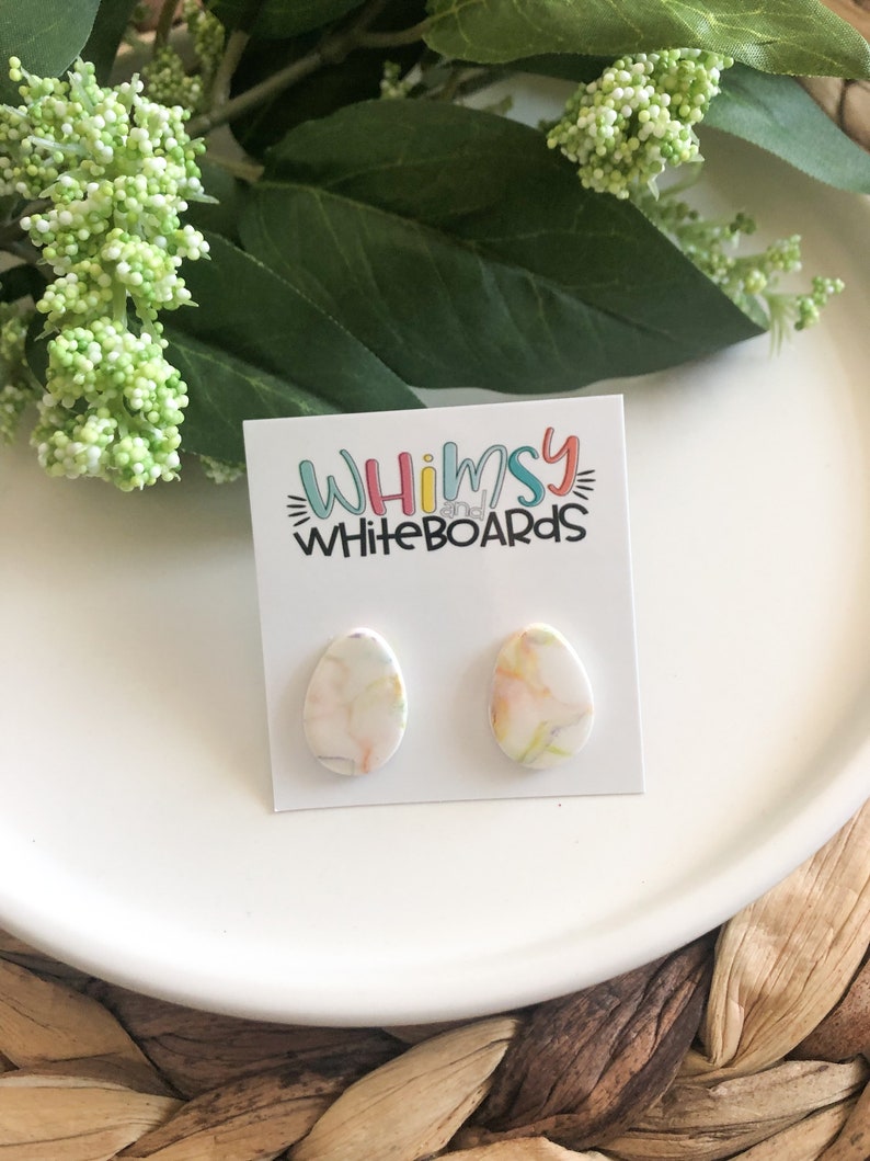May include: A pair of white oval-shaped earrings with a marbled design in shades of pink, yellow, and green. The earrings are displayed on a white card with the text "Whimsy Whiteboards".