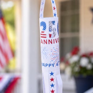 May include: A white banner with a design of red, white, and blue stars and stripes, celebrating the 25th anniversary and Happy 4th. The banner is hanging outdoors, with a flag and flowers in the background.