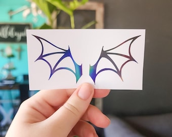 May include: A white card with a black iridescent sticker of two wings. The wings are symmetrical and have a pointed shape with a webbed design.
