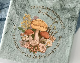 Op de afbeelding: Een lichtgroen T-shirt met een verweerd, tie-dye-effect. Het shirt heeft een afbeelding van paddenstoelen en bloemen met de tekst "THE GRASS WITHERS & THE FLOWERS FADE BUT THE WORD OF THE LORD STANDS FOREVER".