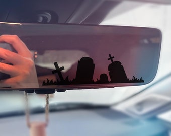 May include: A rearview mirror with a black silhouette of a graveyard with tombstones and crosses. The mirror is hanging from the ceiling of a car.