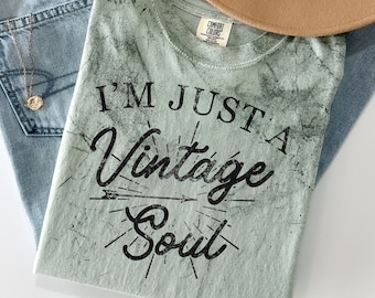 Puede incluir: Una camiseta verde claro con un aspecto desgastado y el texto "I'm Just A Vintage Soul" impreso en negro. La camiseta tiene un aspecto descolorido y vintage y presenta un pequeño diseño de flecha y rayos de sol.