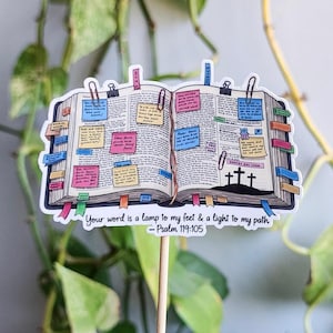 May include: A sticker depicting an open book adorned with colourful sticky notes, clips, and bookmarks. The text "Your word is a lamp to my feet & a light to my path - Psalm 119:105" is at the bottom. Three crosses are drawn on the right page.