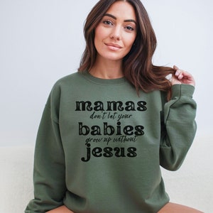 May include: Olive green sweatshirt with the text "mamas don't let your babies grow up without Jesus" in a black, bold font. The sweatshirt is a crew neck style and has long sleeves. The image shows a person wearing the sweatshirt.