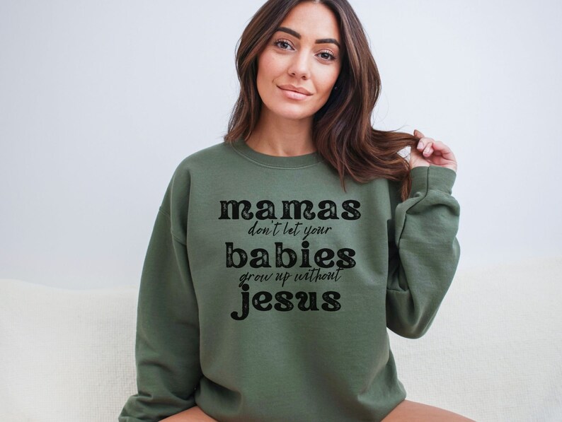 May include: Olive green sweatshirt with the text "mamas don't let your babies grow up without Jesus" in a black, bold font. The sweatshirt is a crew neck style and has long sleeves. The image shows a person wearing the sweatshirt.