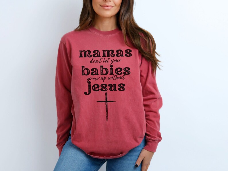 May include: A long-sleeved, red-toned sweatshirt with the text "mamas don't let your babies grow up without jesus" in black lettering. A simple cross is below the text. The sweatshirt is a casual style.