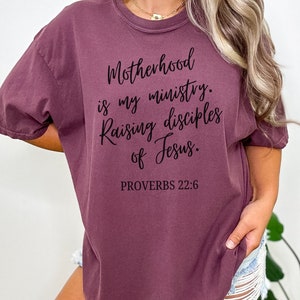 May include: A maroon t-shirt with the text "Motherhood is my ministry. Raising disciples of Jesus. PROVERBS 22:6" printed in black.