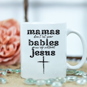 May include: White ceramic mug with the text "mamas don't let your babies grow up without jesus" in black. A black cross is below the text. The mug has a curved handle and is surrounded by a blurred background with a pink flower and decorative glass beads.