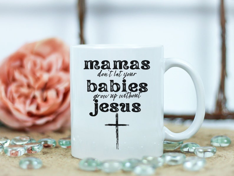 May include: White ceramic mug with the text "mamas don't let your babies grow up without jesus" in black. A black cross is below the text. The mug has a curved handle and is surrounded by a blurred background with a pink flower and decorative glass beads.