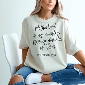 May include: A woman wearing a beige t-shirt with the text "Motherhood is my ministry. Raising disciples of Jesus. Proverbs 22:6" printed in black.