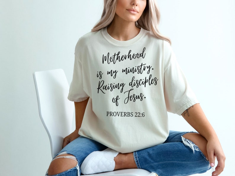 May include: A woman wearing a beige t-shirt with the text "Motherhood is my ministry. Raising disciples of Jesus. Proverbs 22:6" printed in black.