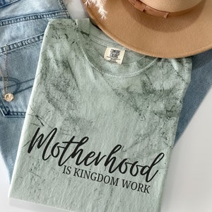 May include: A light green t-shirt with a distressed, marbled pattern. The shirt has black text that reads "Motherhood is Kingdom Work."