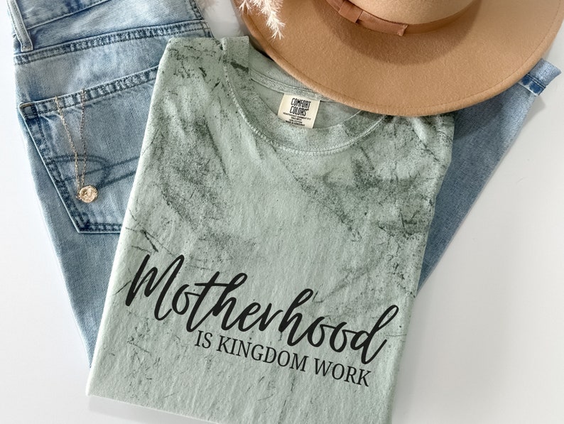 May include: A light green t-shirt with a distressed, marbled pattern. The shirt has black text that reads "Motherhood is Kingdom Work."