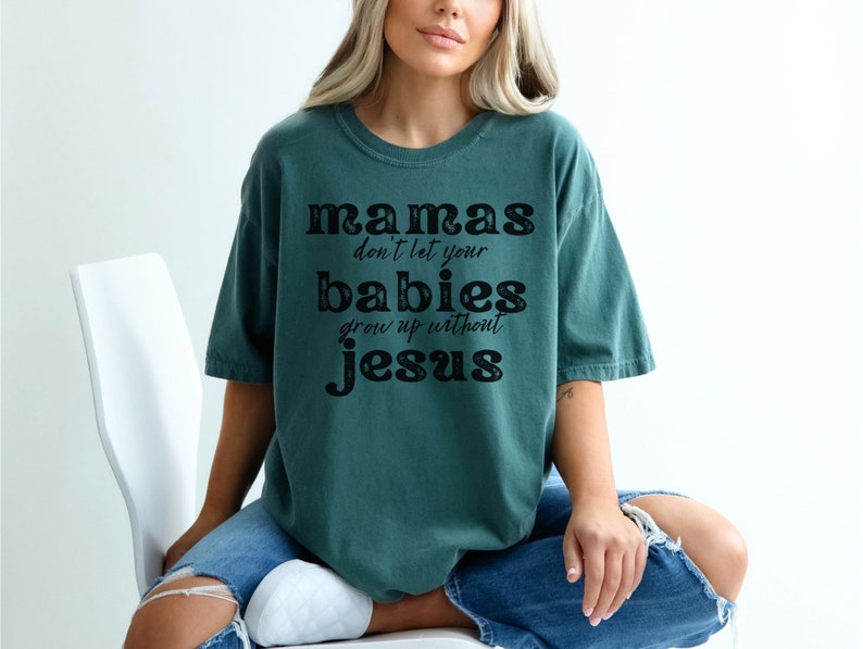 May include: Teal-colored t-shirt with the text "mamas don't let your babies grow up without jesus" in black lettering. The shirt has a relaxed fit and short sleeves. The text is in a bold, stylized font.