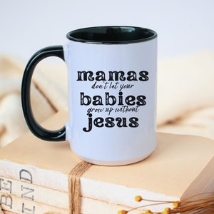 May include: White ceramic mug with a black handle and rim. The mug features the text "mamas don't let your babies grow up without jesus" in black lettering. The mug is sitting on a stack of books.