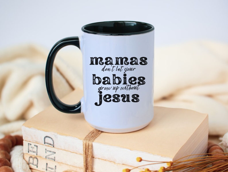 May include: White ceramic mug with a black handle and rim. The mug features the text "mamas don't let your babies grow up without jesus" in black lettering. The mug is sitting on a stack of books.