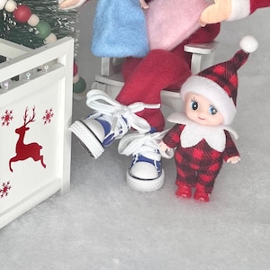 May include: A small elf doll wearing a red and black plaid outfit with a white hat. The elf is holding a red and white candy cane. The elf is sitting on a white wooden chair.
