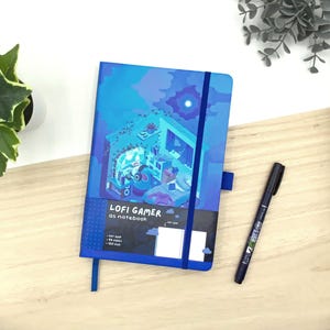 Peut inclure: Un cahier bleu avec l'inscription "LOFI GAMER as notebook" sur la couverture. La couverture présente une illustration d'une pièce avec une personne portant un casque. Un stylo noir est à côté du cahier.