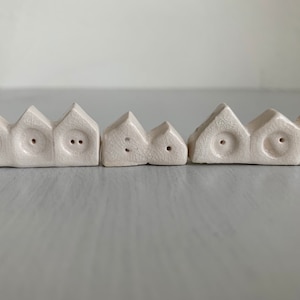 Könnte beinhalten: Drei cremefarbene, hausförmige Keramikobjekte. Jedes hat ein dreieckiges Dach und kreisförmige Löcher, die Fenstern oder Augen ähneln. Die Objekte variieren in der Größe und haben eine strukturierte, rissige Oberfläche.
