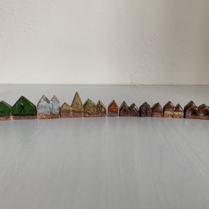 Könnte beinhalten: Eine Reihe von Miniatur-Keramikhäusern in verschiedenen Formen und Farben. Die Häuser weisen hauptsächlich grüne, braune und beige Farbtöne auf, einige mit strukturierten Details. Die Gesamtästhetik ist rustikal und handgefertigt.