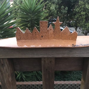 Könnte beinhalten: Handgefertigte Silhouette der Skyline von San Francisco aus Kork. Die Stadtlandschaft zeigt ikonische Gebäude und einen Stern, montiert auf einer hellbraunen ovalen Basis. Die Worte "San Francisco" sind handschriftlich auf der Basis.