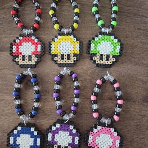 May include: Six colorful mushroom-shaped beaded necklaces. Each necklace features a different color mushroom pendant: red, yellow, green, blue, purple, and pink. The beads on the necklaces are black, clear, and the same color as the mushroom.