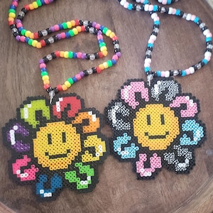 May include: Two beaded necklaces with colorful flower charms. The flowers are pixelated and feature a smiley face in the center. One necklace is rainbow colored and the other is black, white, and blue.