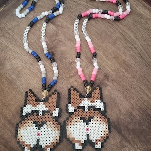 May include: Two beaded necklaces with pixelated dog charms. The necklaces are made with white, gray, blue, pink, and black beads. The necklaces spell out "RAVE" and "BOOTY".