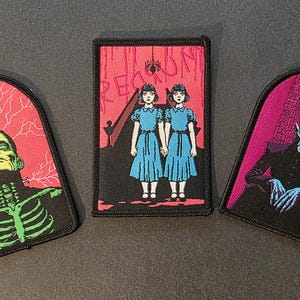 May include: Three embroidered patches with horror movie themes. One features Frankenstein's monster, another shows the Grady twins from The Shining, and the third depicts a vampire. Each patch has a black border and a unique design.